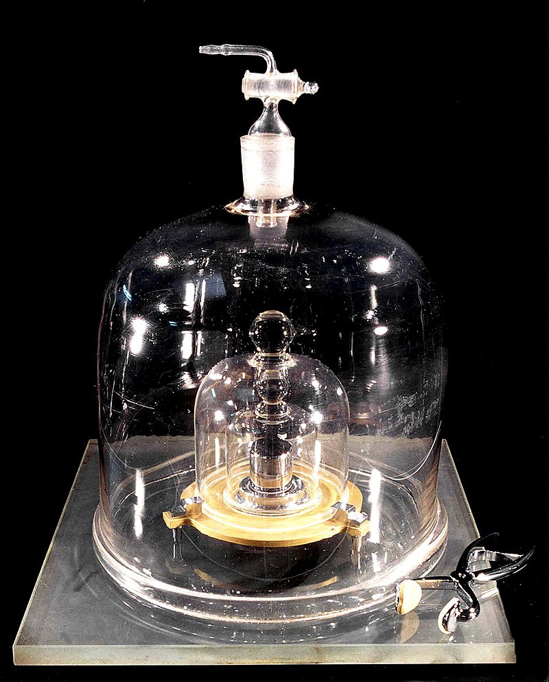 A shiny metal cylinder sits on a glass base under two nested glass bell jars. A pair of tongs is placed beside the base. 