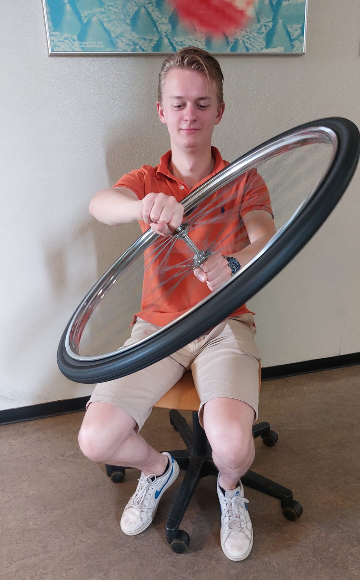 A male student with blonde hair is sitting on a swivel chair. He is holding the axle of a bicycle wheel in front of him, at a 45 degree angle. 