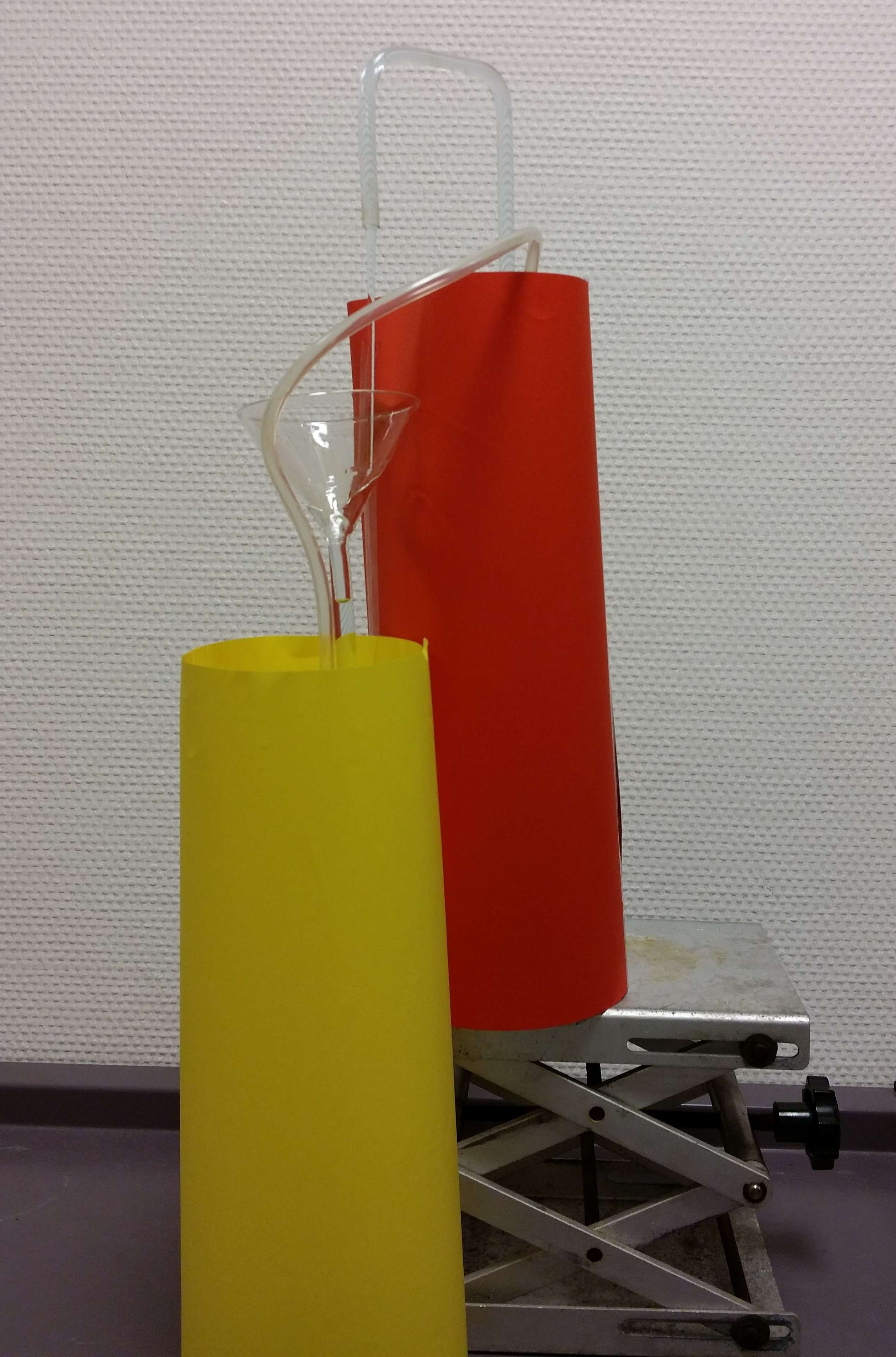 A laboratory setup featuring two large cylindrical containers wrapped in colored paper—one yellow (left) and one red (right)—connected by a transparent plastic tube. A glass funnel is inserted into the yellow container, with the tube extending from the funnel to the red container. The red container is elevated on a metallic lab jack with a scissor-lift mechanism.